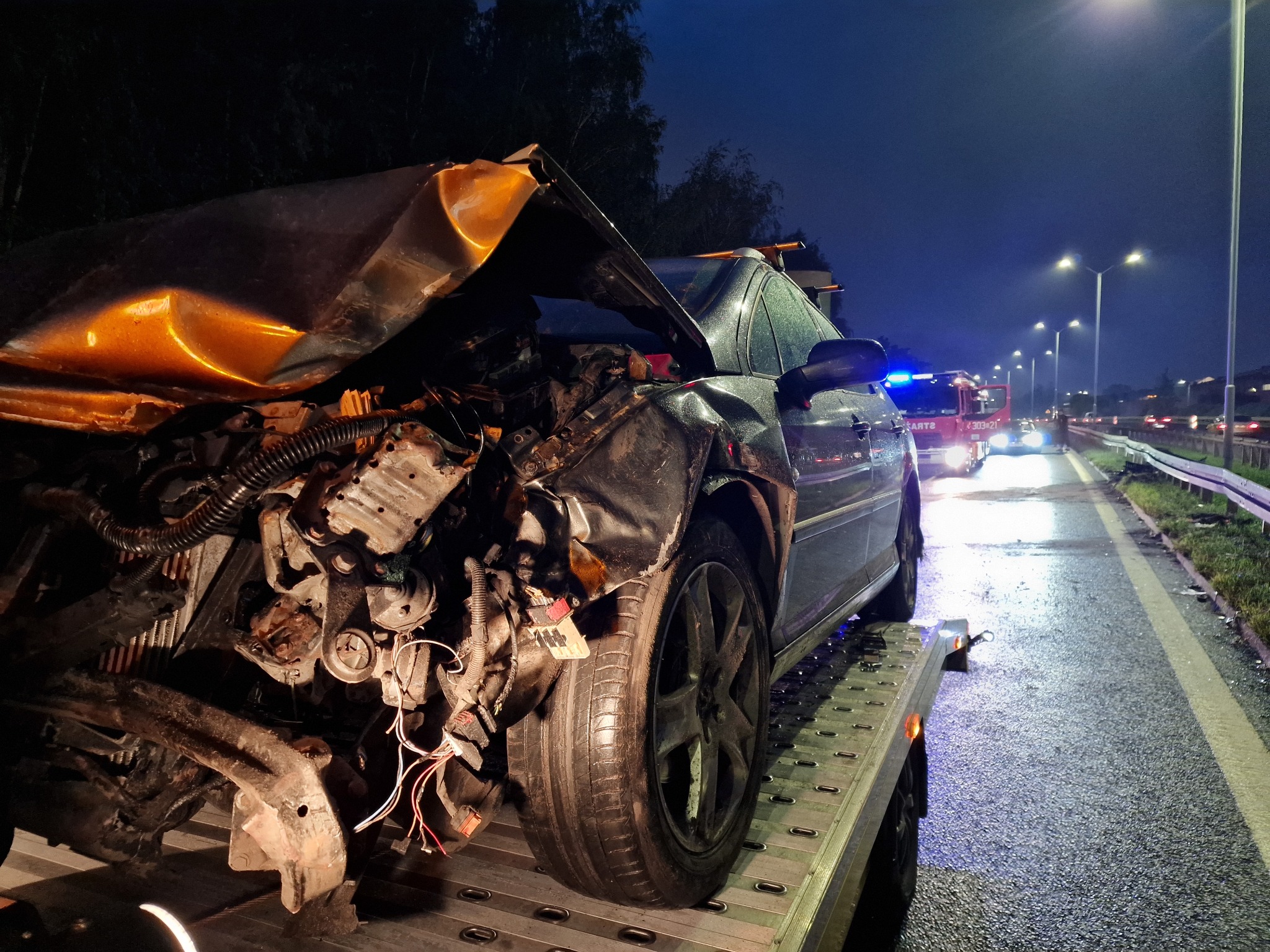 Uszkodzony samochód osobowy z widocznymi zniszczeniami karoserii i silnika, umieszczony na platformie lawety w nocnych warunkach, z włączonymi światłami pojazdu straży pożarnej w tle.