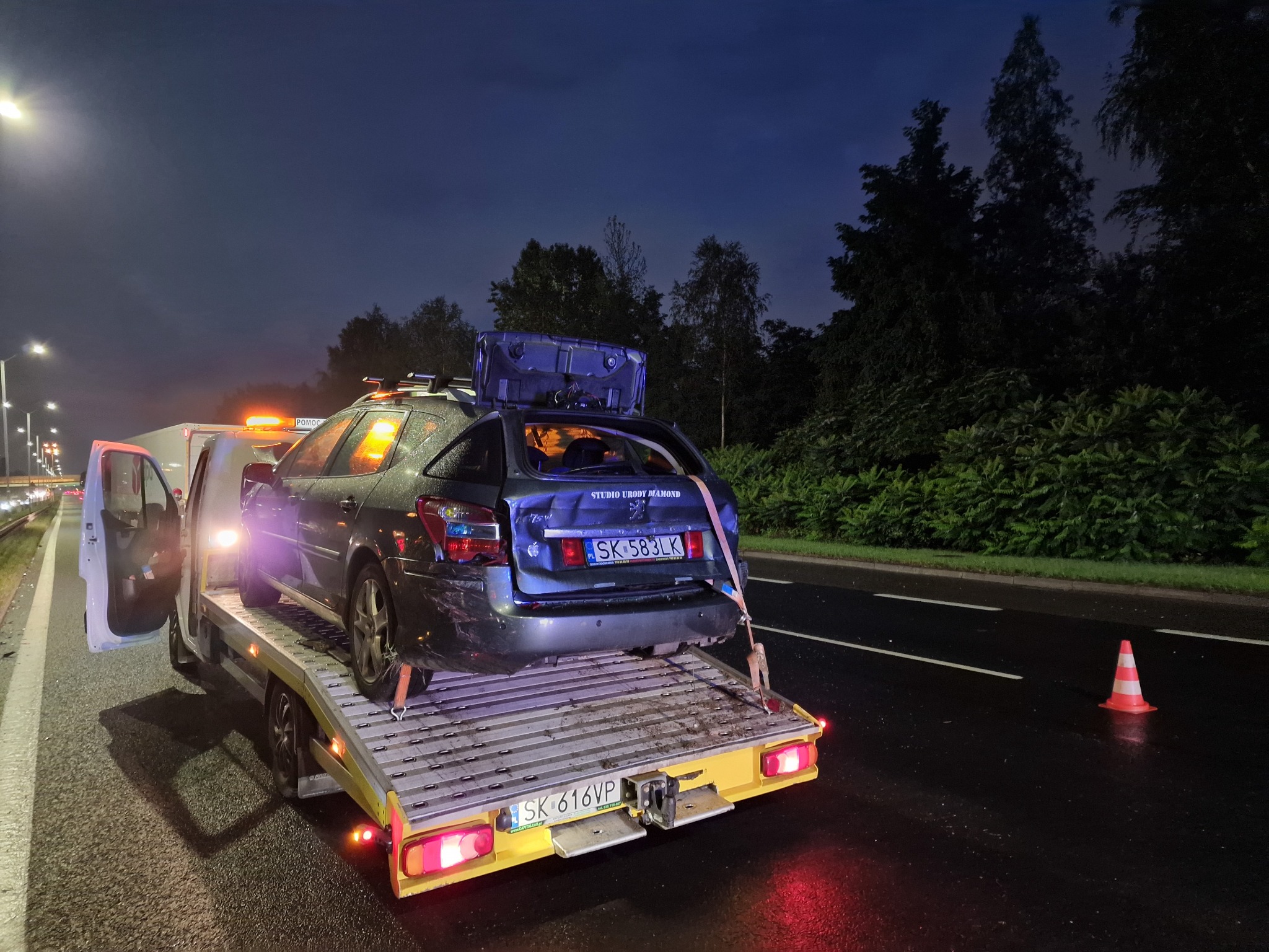 Uszkodzony samochód osobowy z otwartym bagażnikiem, z bagażem na dachu, zabezpieczony pasami transportowymi na platformie lawety w nocnych warunkach, na mokrej nawierzchni drogi, z widocznym...