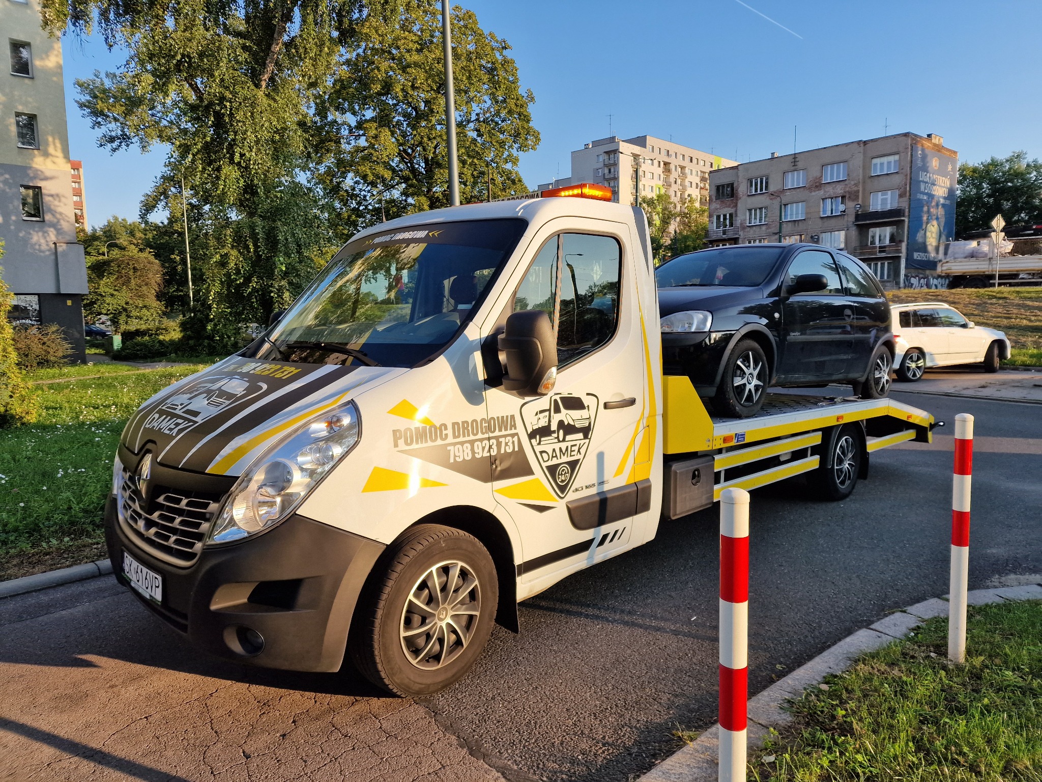 Biała laweta Renault Master z żółtymi akcentami i logo firmy 'DAMEK' przewozi czarny samochód osobowy na tle miejskiego krajobrazu z budynkami i zielenią.