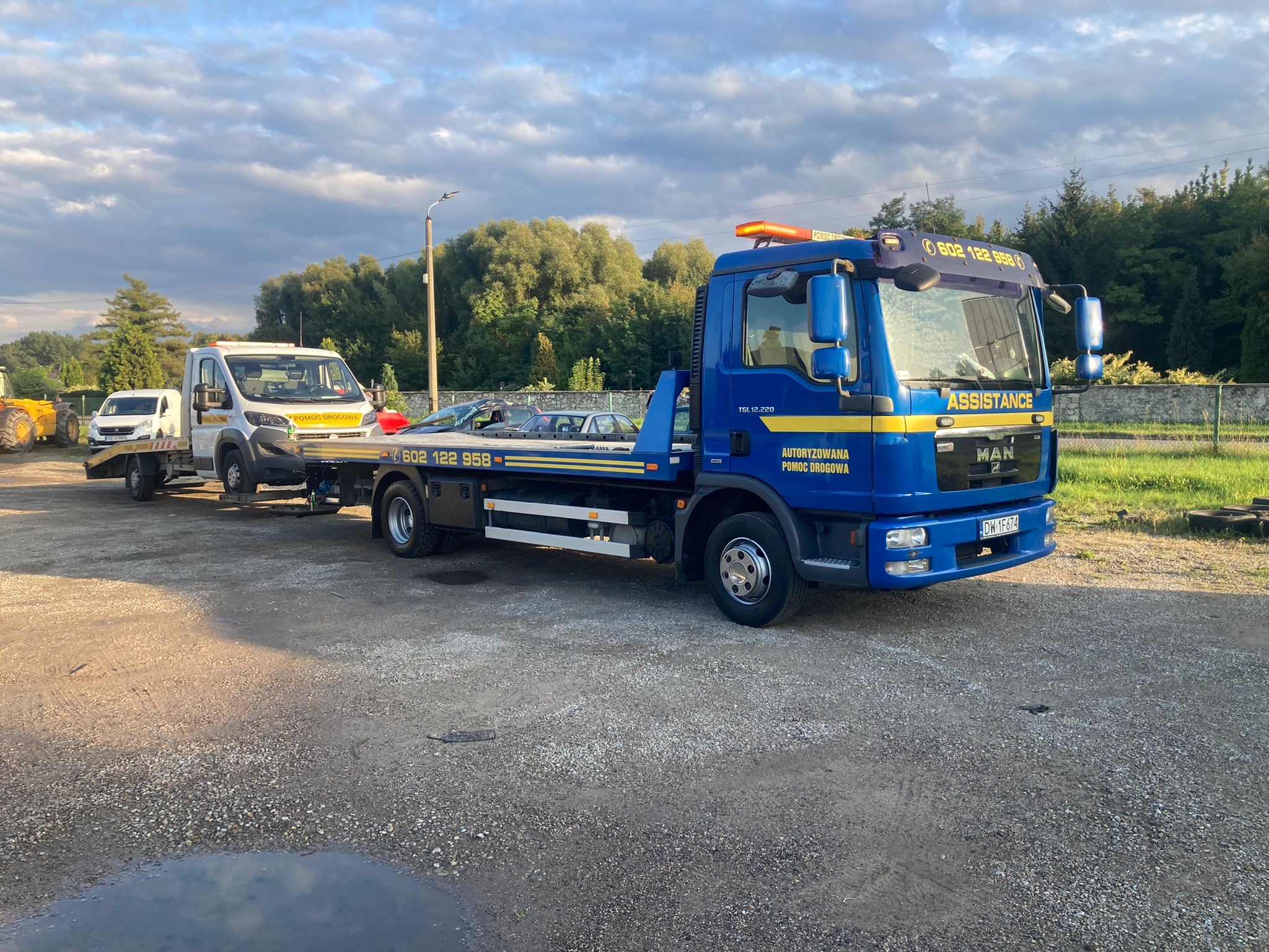 Niebieska laweta MAN z żółtymi pasami Assistance holuje uszkodzony biały samochód dostawczy na platformie, dodatkowo za lawetą stoi biały bus na lawecie, widok z boku na tle zieleni i pochmurnego...