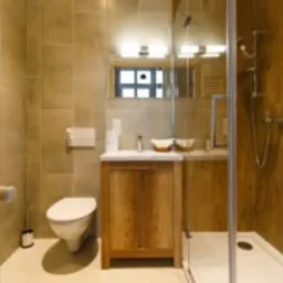 Nowoczesna łazienka z kabiną prysznicową typu walk-in, wiszącą toaletą i szafką pod umywalką z drewnianymi frontami. Beżowe płytki na ścianach i podłodze, lustro z oświetleniem nad umywalką.