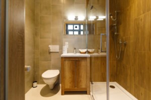 Nowoczesna łazienka z kabiną prysznicową typu walk-in, wiszącą toaletą i szafką pod umywalką z drewnianymi frontami. Beżowe płytki na ścianach i podłodze, lustro z oświetleniem nad umywalką.