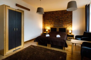 Sypialnia z łóżkiem małżeńskim z brązową narzutą i białymi ręcznikami, drewnianą szafą z czarnymi frontami, dywanem w kolorze czekolady, ceglaną ścianą za wezgłowiem i dwoma lampami nocnymi.