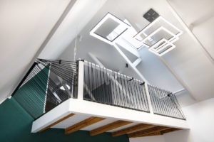 Nowoczesna antresola z czarną balustradą na białej platformie, widok z dołu na skośny sufit z kwadratowymi lampami LED.