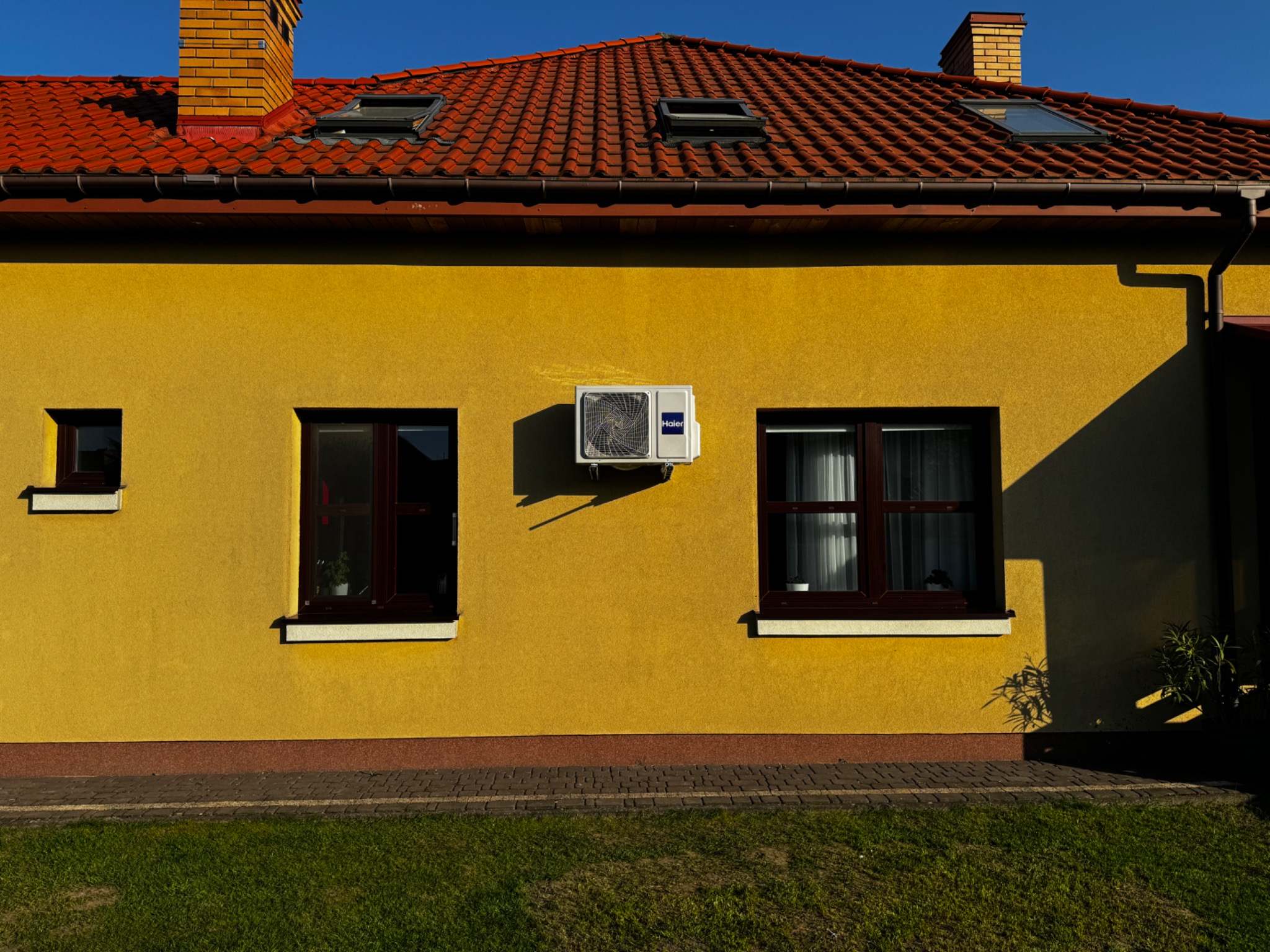 Żółta ściana domu z zamontowanym białym klimatyzatorem Haier, widoczne okna z brązowymi ramami, czerwony dach i zielony trawnik.