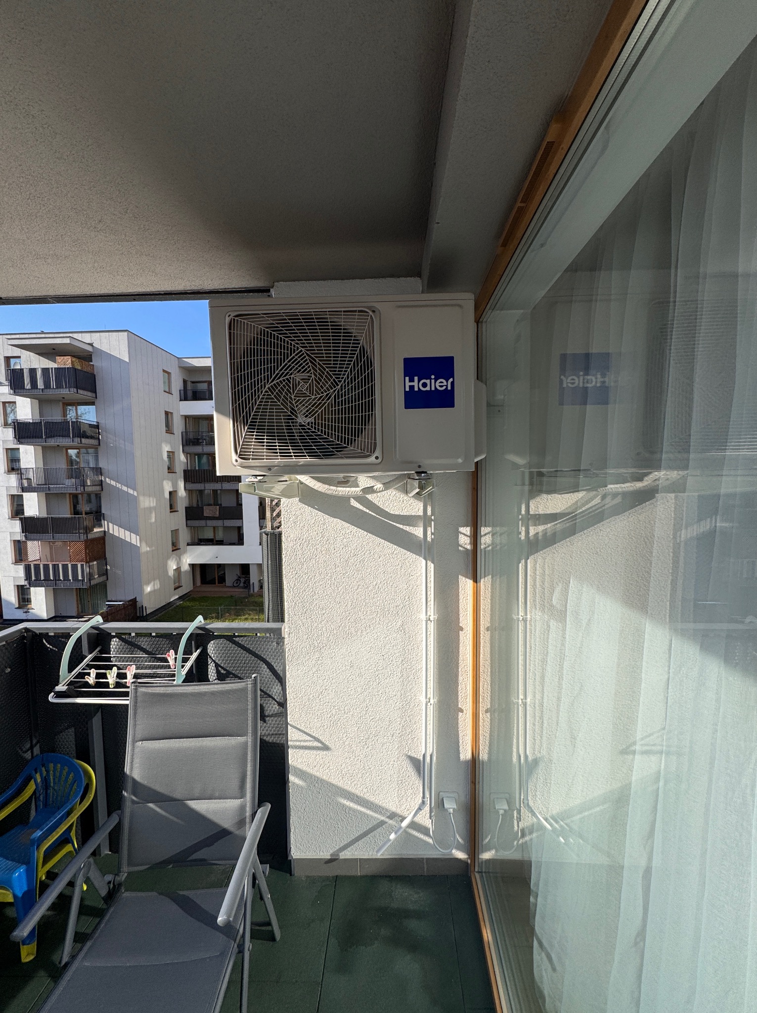 Zewnętrzna jednostka klimatyzacji Haier zamontowana na balkonie mieszkania w bloku, widoczne kable i rurki prowadzące do wnętrza.