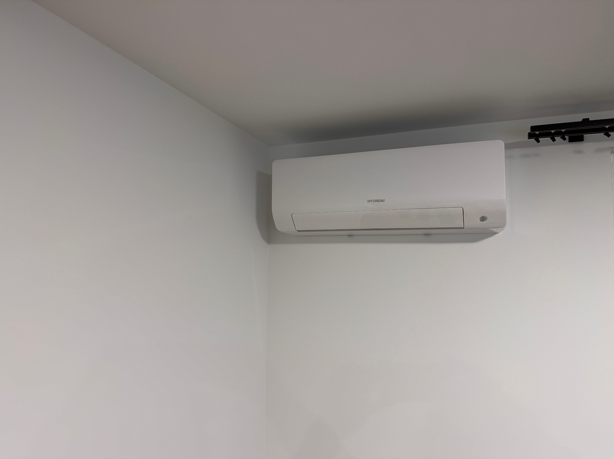 Biały klimatyzator marki Hyundai zamontowany na ścianie w jasnym pomieszczeniu, widoczne szyny oświetleniowe na suficie.