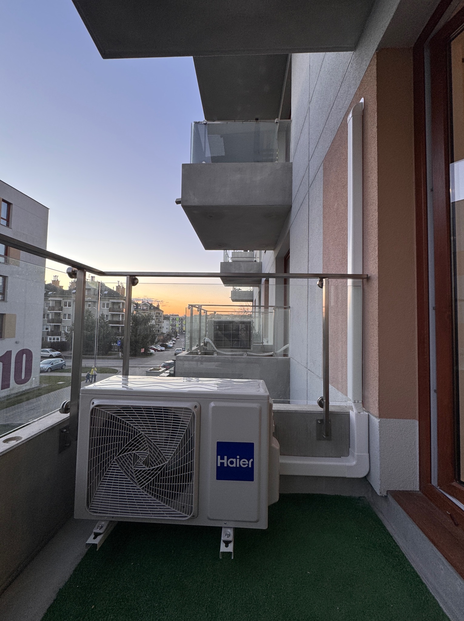 Biała jednostka zewnętrzna klimatyzatora Haier zamontowana na balkonie apartamentu z widokiem na zachodzące słońce.