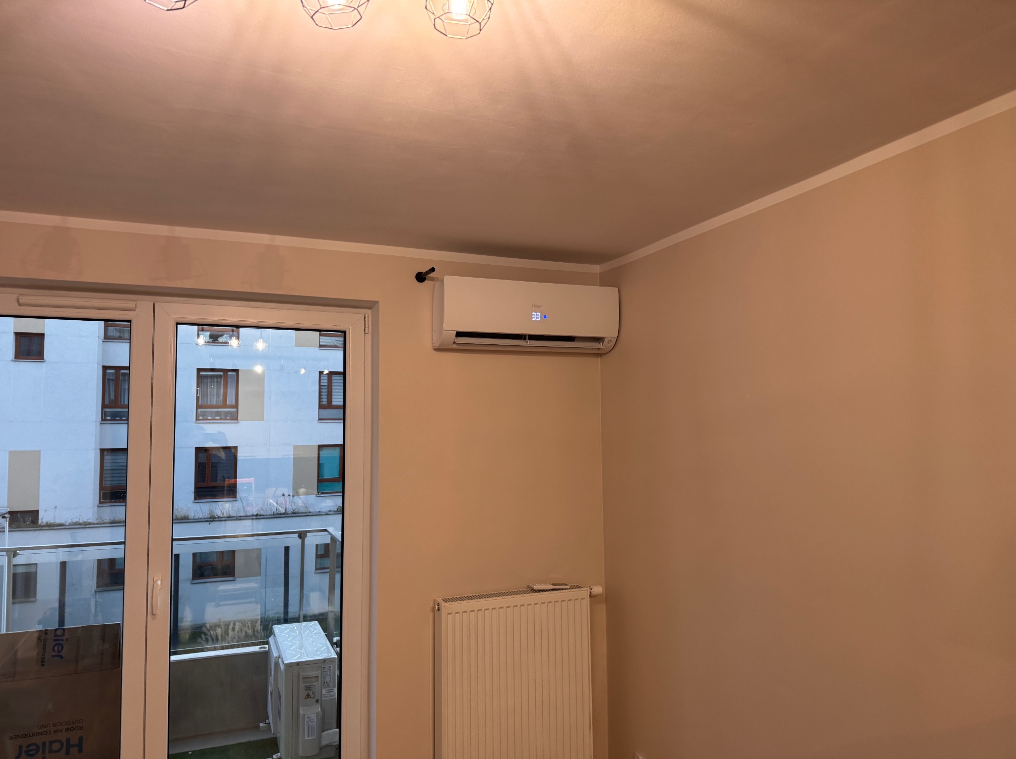 Zamontowany klimatyzator w jasnym pokoju, widoczny wyświetlacz z temperaturą 33 stopnie, na balkonie jednostka zewnętrzna w kartonie.
