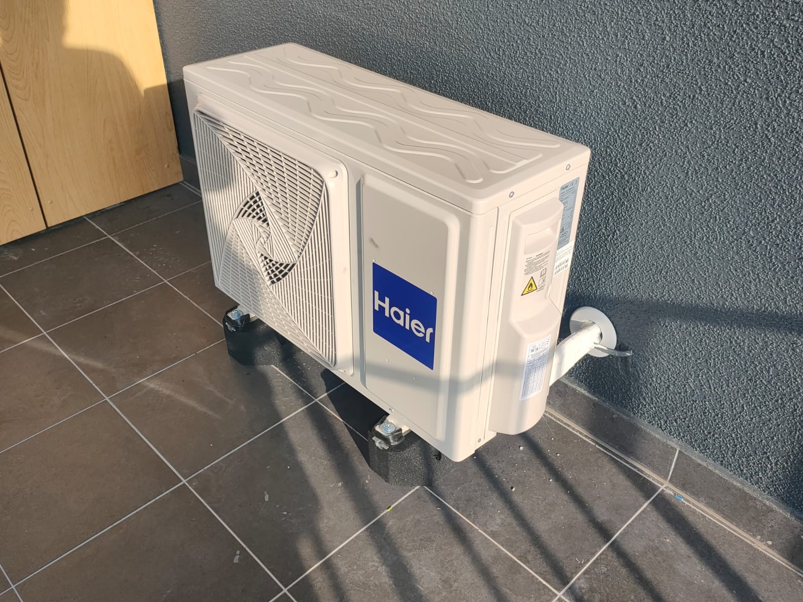 Zewnętrzna jednostka klimatyzacji Haier zamontowana na czarnych podkładkach antywibracyjnych na szarym balkonie z płytkami, rury odprowadzające ciepło przymocowane do ściany.