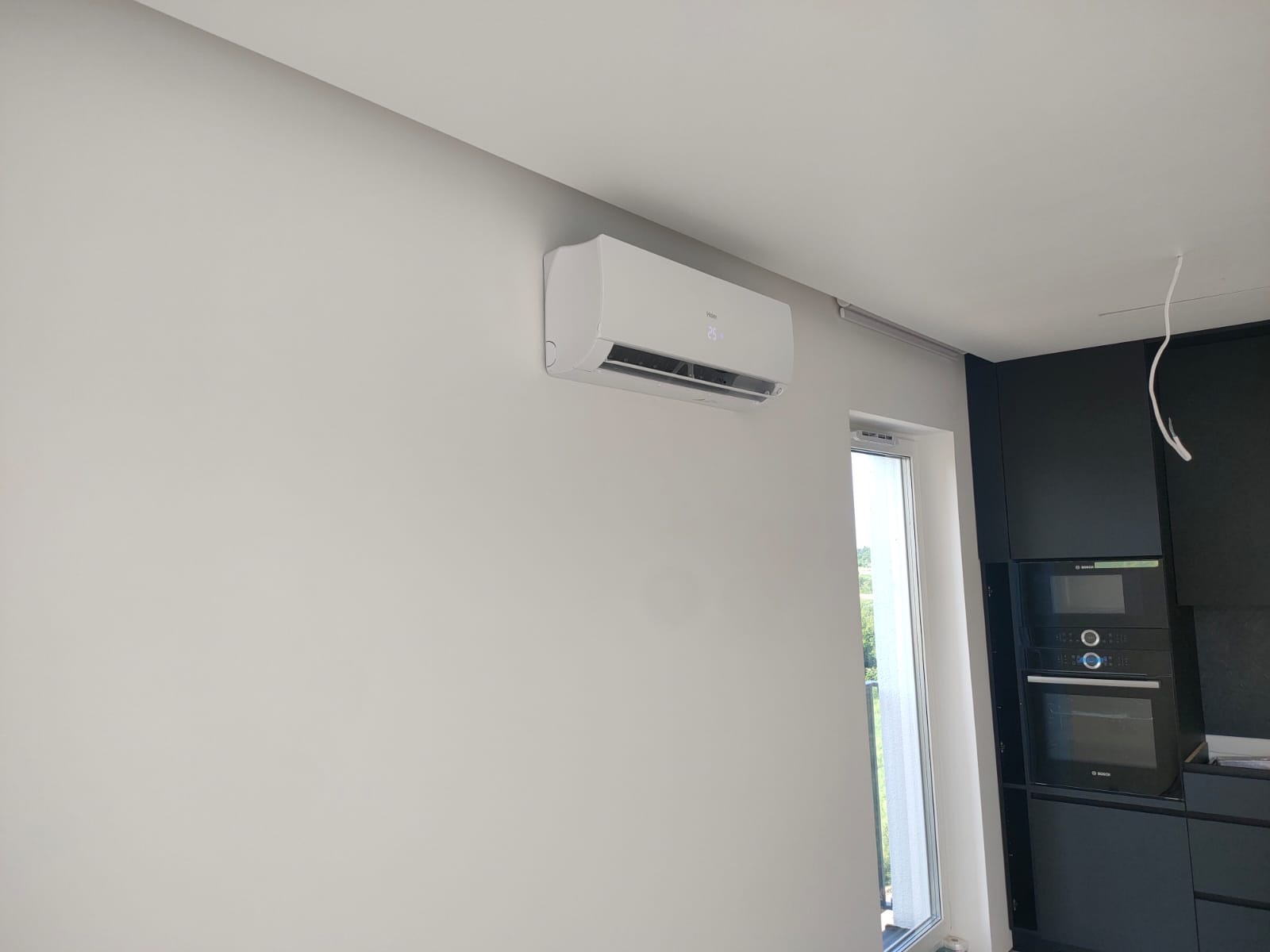 Biały klimatyzator Haier zamontowany na ścianie w nowoczesnym wnętrzu, obok okna i czarnych szafek kuchennych.