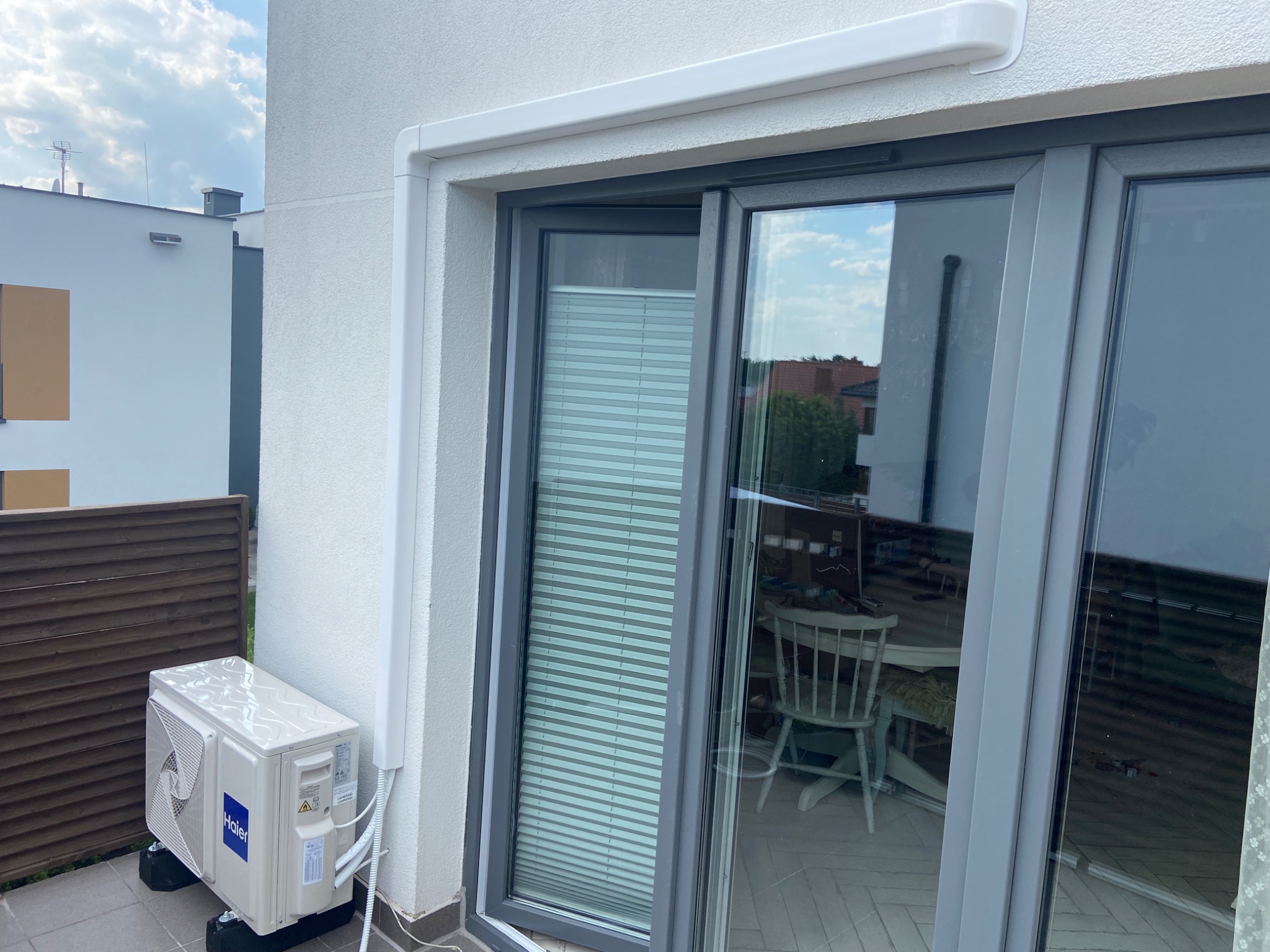 Biała jednostka zewnętrzna klimatyzacji Haier, zamontowana na balkonie z szarą podłogą, widoczna zza szarych drzwi balkonowych z żaluzjami wewnętrznymi.