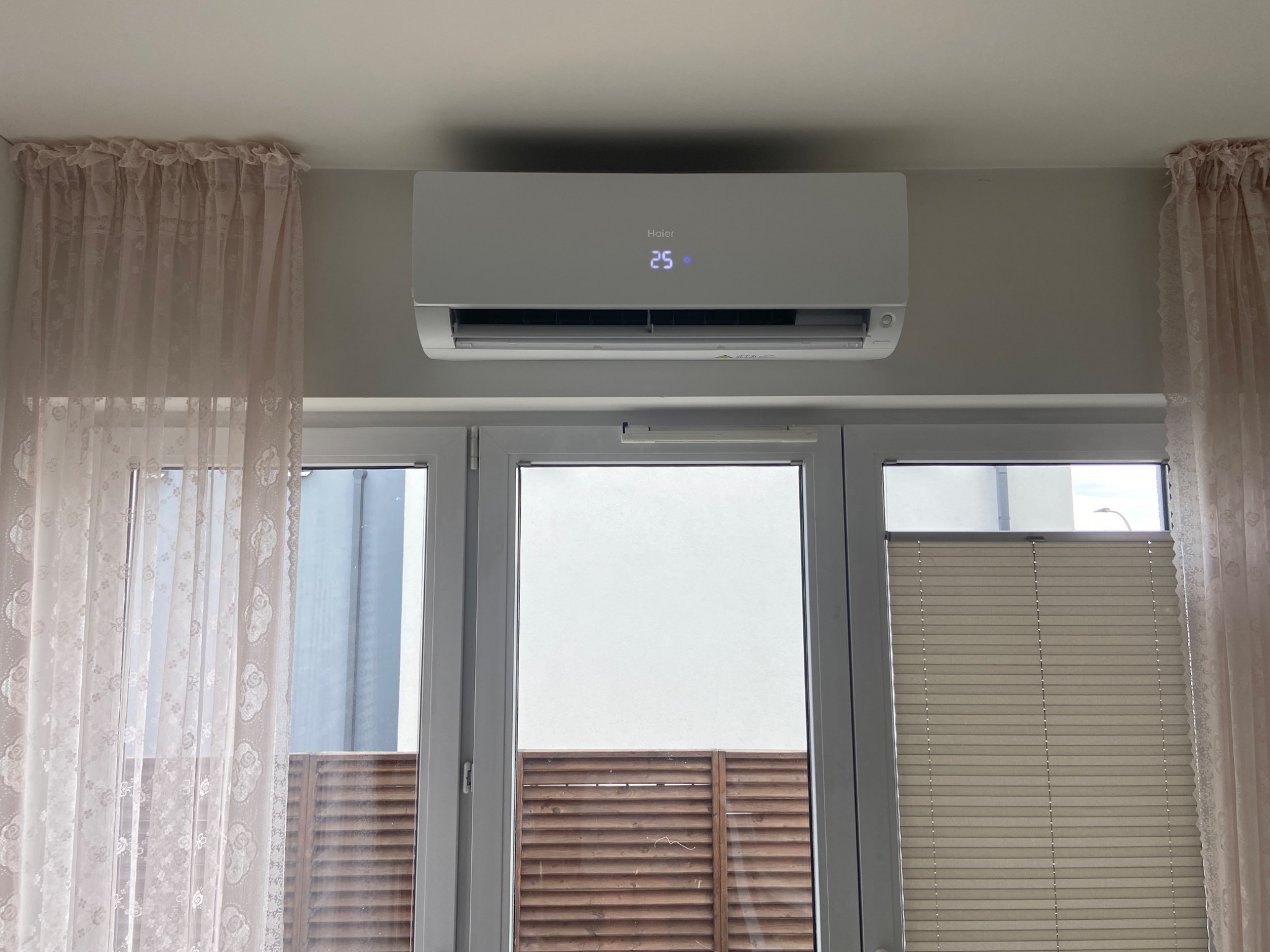 Biały klimatyzator Haier z wyświetlaczem temperatury 25 stopni Celsjusza, zamontowany nad oknem z zasłonami i żaluzjami.