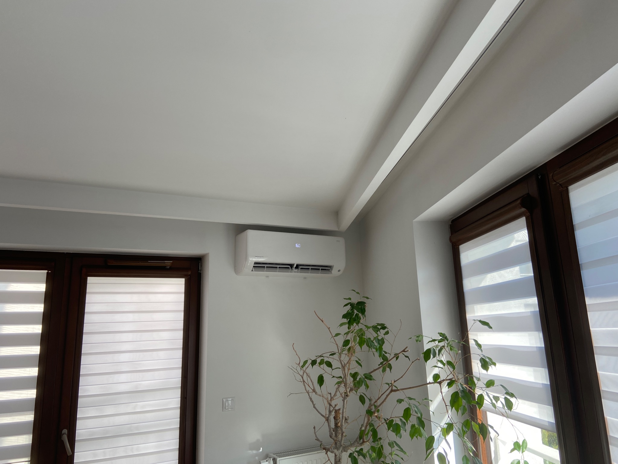 Biała jednostka klimatyzacji zamontowana na ścianie w jasnym pomieszczeniu, widoczna z boku, z rośliną doniczkową w tle i oknami z żaluzjami dzień-noc.