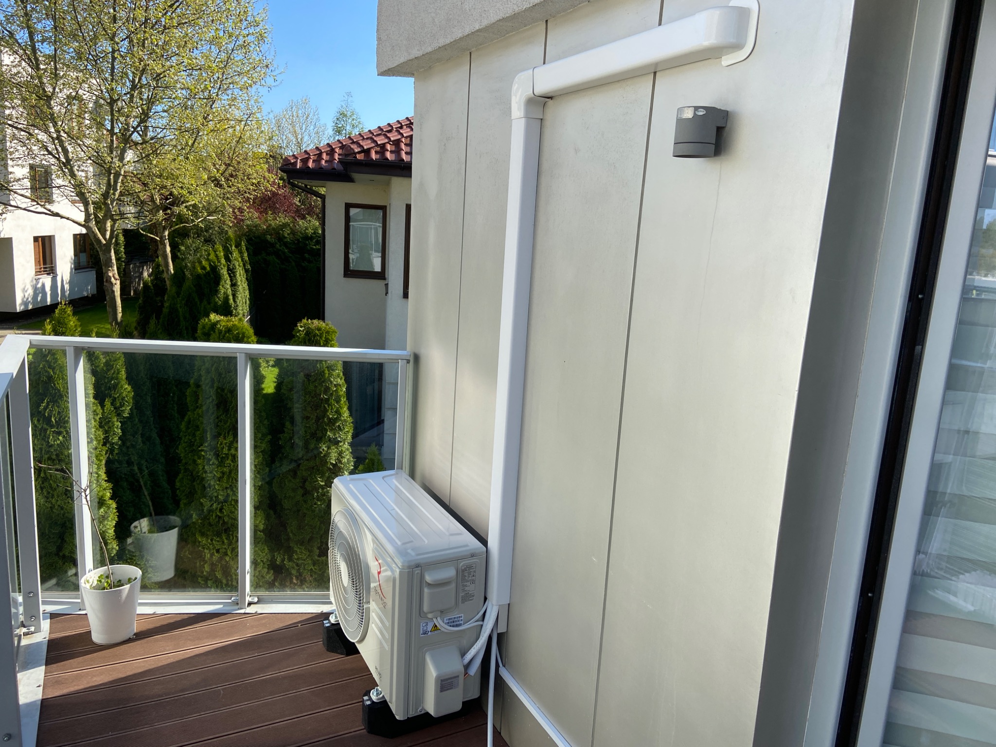 Jednostka zewnętrzna klimatyzacji zamontowana na balkonie z drewnianą podłogą, widoczne białe rury odprowadzające, w tle zieleń i budynek.