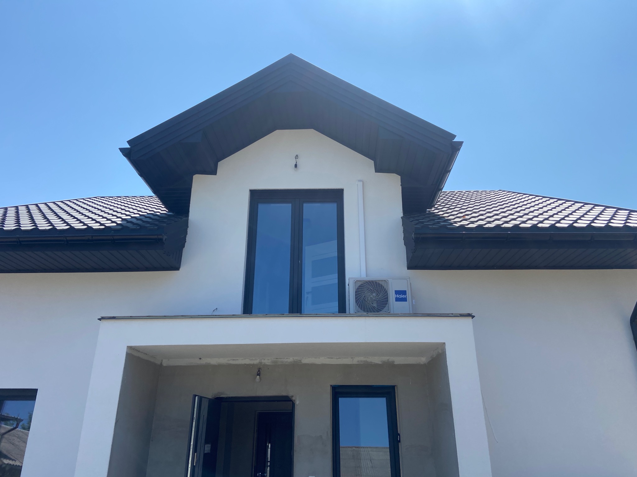 Biały dom jednorodzinny z zamontowaną jednostką zewnętrzną klimatyzacji Haier, widok z dołu w słoneczny dzień.