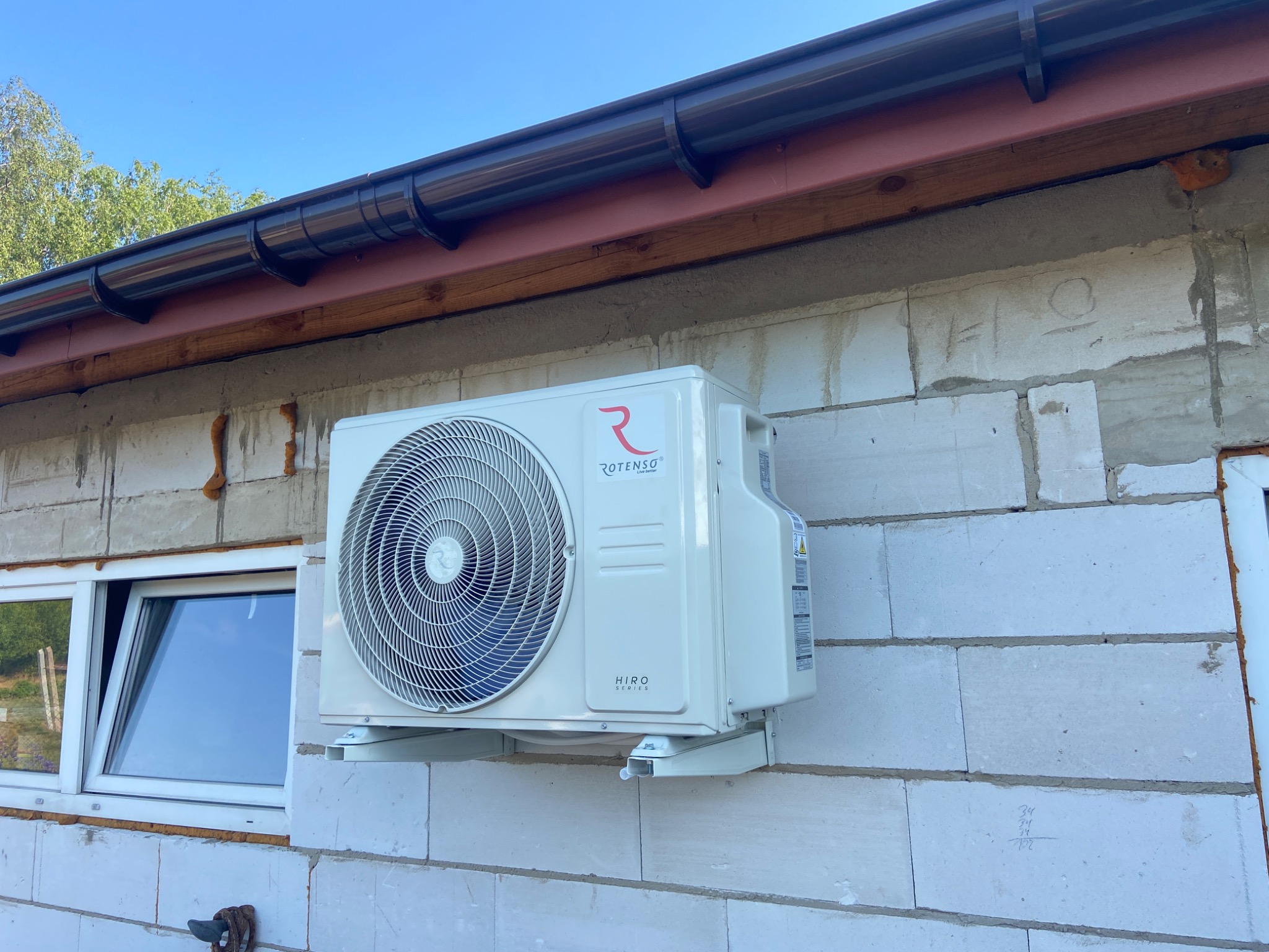 Zewnętrzna jednostka klimatyzacji Rotenso Hiro Series zamontowana na ścianie budynku z bloczków betonowych, widoczne okno z białymi ramami i brązowe orynnowanie.