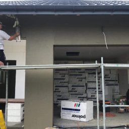 Clean Company - Pracownik na rusztowaniu wyrównuje zaprawę na elewacji budynku w trakcie budowy, widoczne ocieplenie wewnątrz, puszki farby i opakowania materiałów budowlanych.