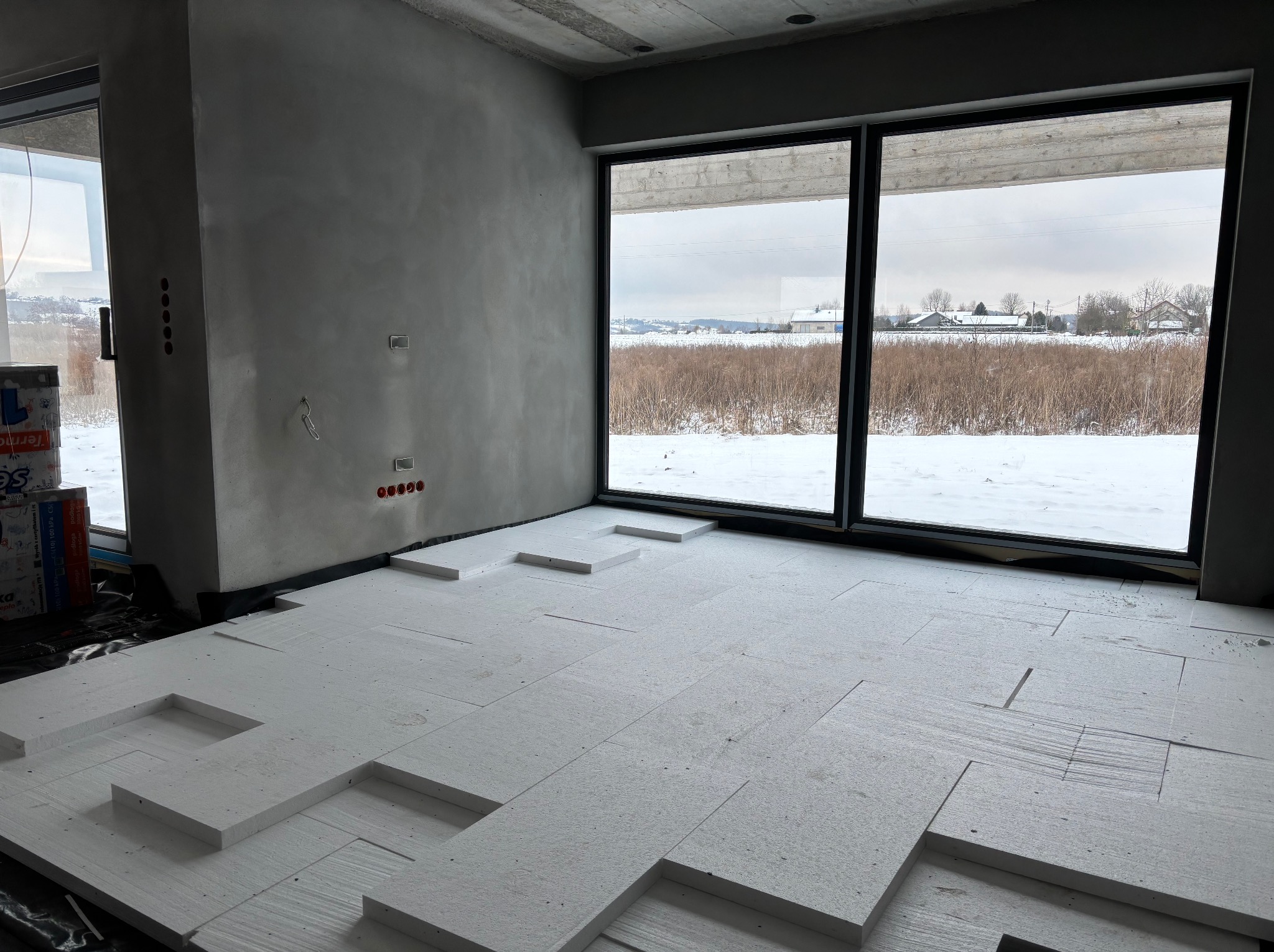 Wnętrze w trakcie prac: białe płyty izolacyjne układane na podłodze, surowa ściana z widocznymi puszkami elektrycznymi, duże okno z widokiem na zimowy krajobraz.