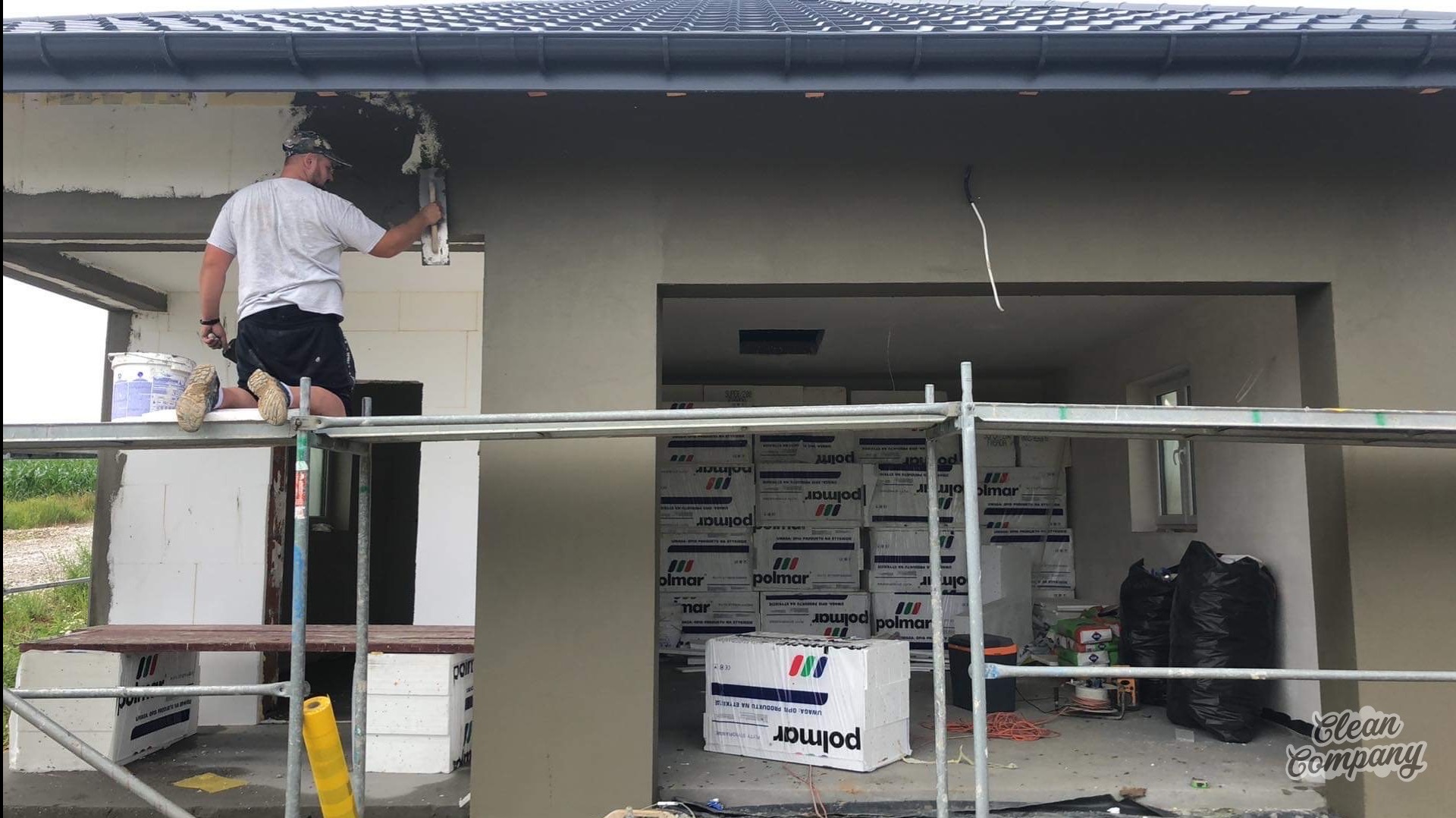 Pracownik na rusztowaniu wyrównuje zaprawę na elewacji budynku w trakcie budowy, widoczne ocieplenie wewnątrz, puszki farby i opakowania materiałów budowlanych.