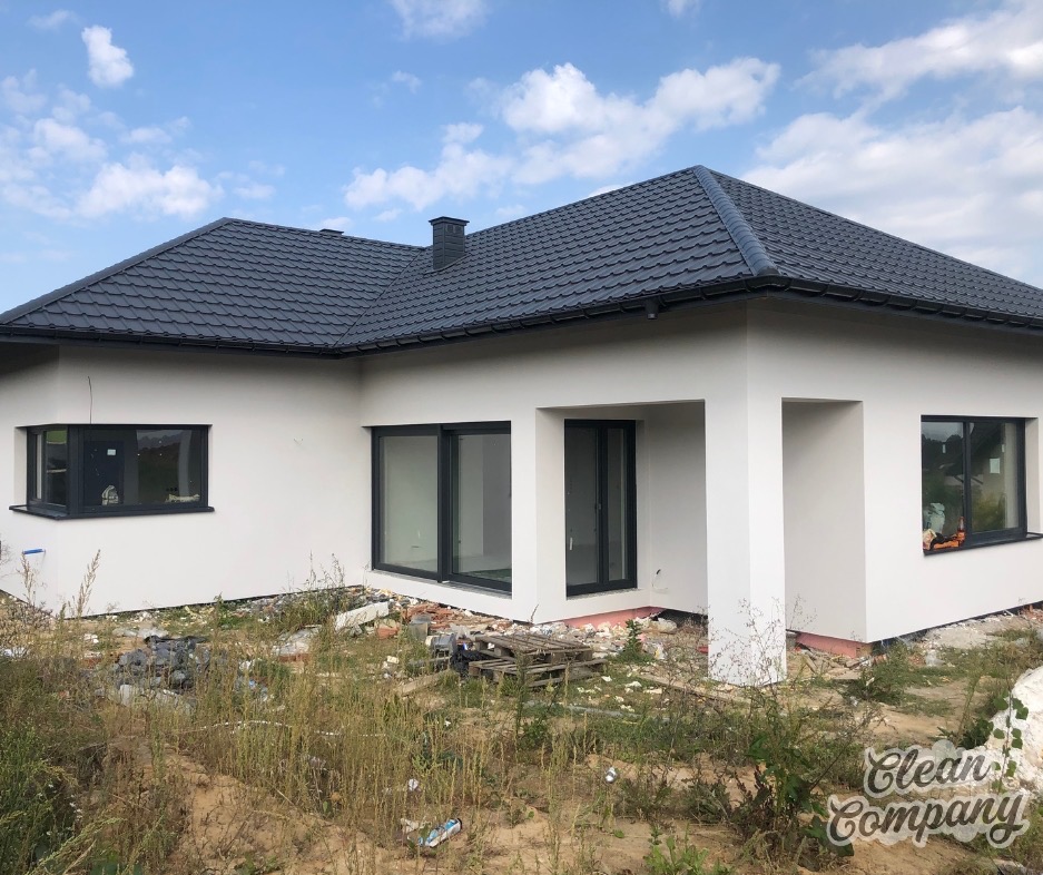 Nowoczesny dom jednorodzinny z grafitowym dachem i jasną elewacją, widok z boku na niezagospodarowany teren z resztkami gruzu i chwastami, logo firmy sprzątającej w prawym dolnym rogu.