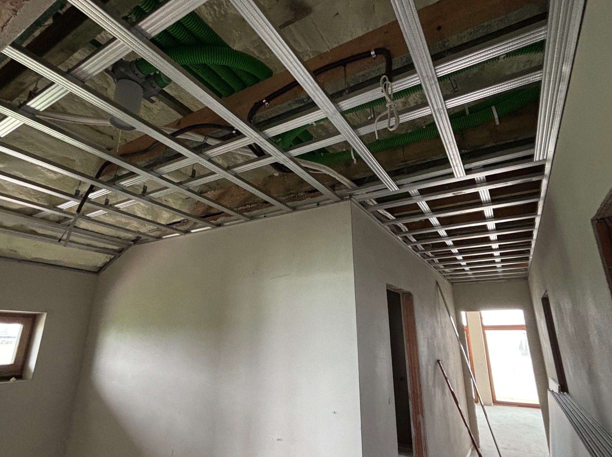 Sufit podwieszany z metalową konstrukcją, widoczne instalacje w zielonych peszlach. Ściany przygotowane do wykończenia, surowy stan wnętrza. Ujęcie z dołu.