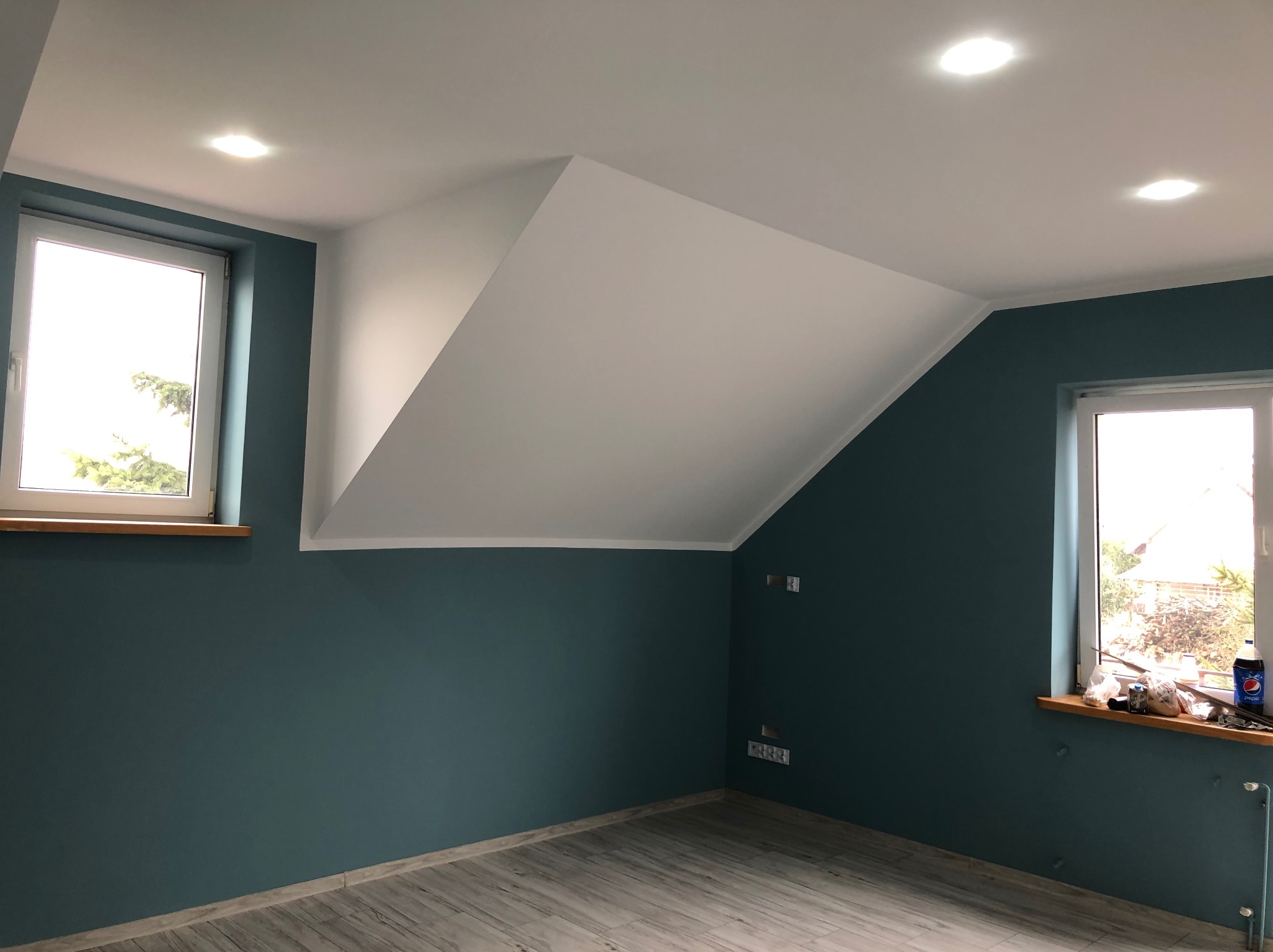 Pomieszczenie na poddaszu po remoncie: ściany w kolorze morskim, biały sufit ze skosem, okna z drewnianymi parapetami, panele podłogowe imitujące drewno.
