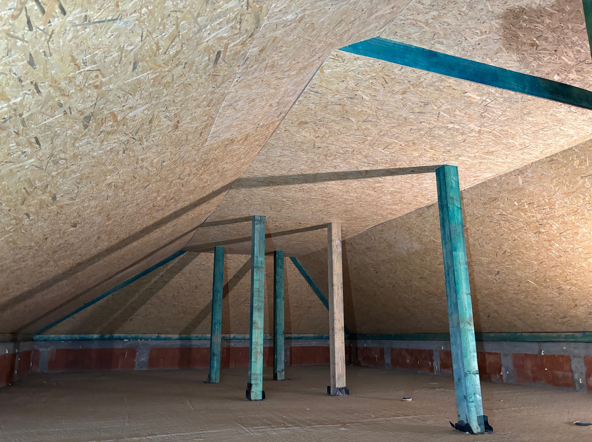 Poddasze z płyt OSB, widoczne drewniane, pomalowane na zielono słupy podtrzymujące konstrukcję dachu. Na podłodze wylewka. Ceglana ściana w tle.