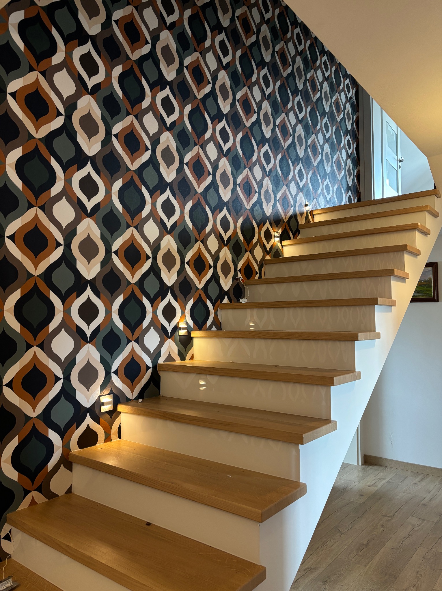 Schody z drewnianymi stopniami i wzorzystą tapetą w tle, oświetlone punktowymi lampkami. Nowoczesny design wnętrza z akcentem retro.