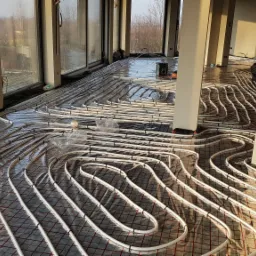 Białe rury grzewcze układane na folii z siatką w nowym budynku z dużymi oknami, widoczne narzędzia i elementy instalacji.