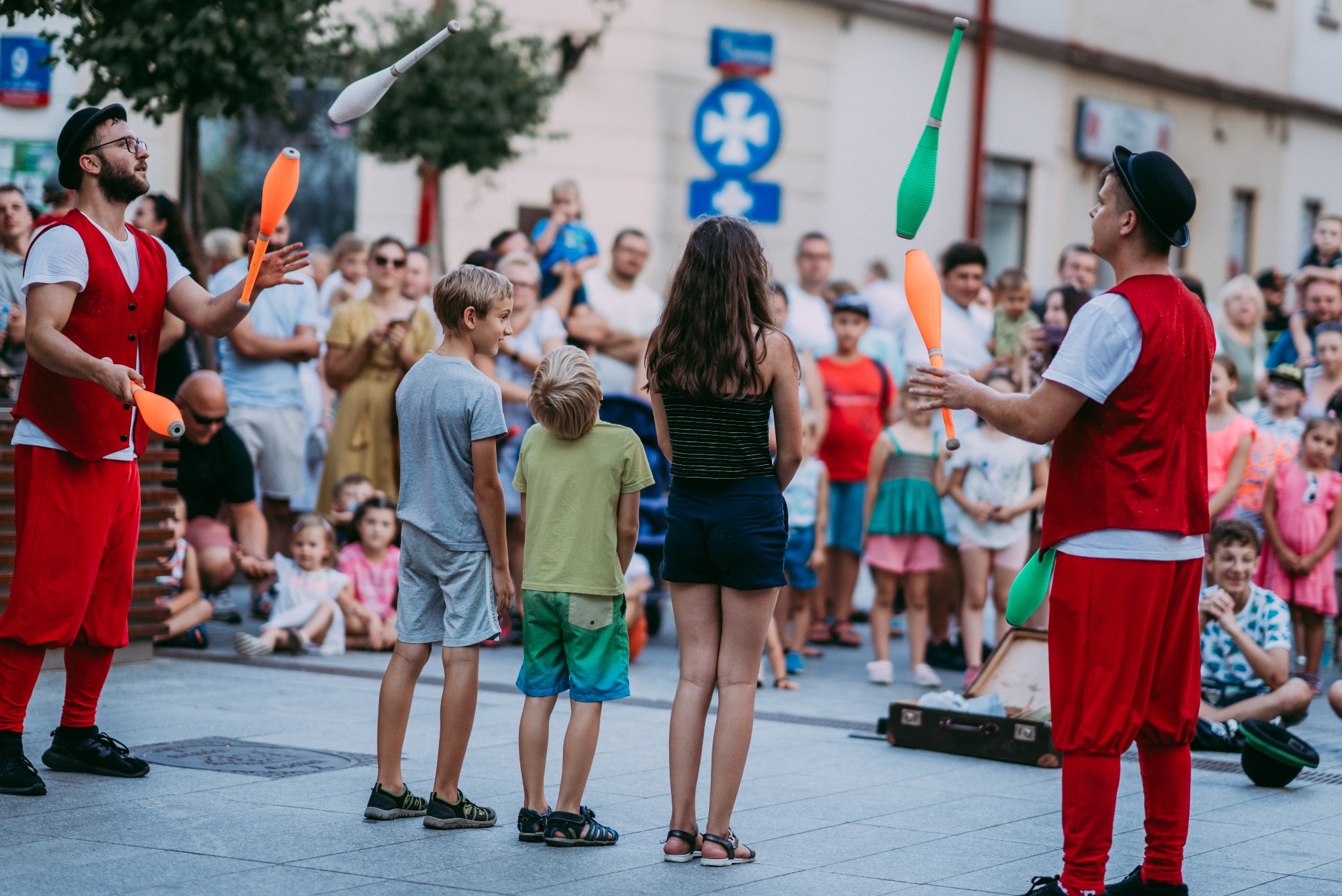 Uliczny pokaz żonglerki maczugami w Rzeszowie, dwóch artystów w czerwonych strojach prezentuje swoje umiejętności przed zgromadzoną publicznością, w tym grupą dzieci stojących najbliżej.