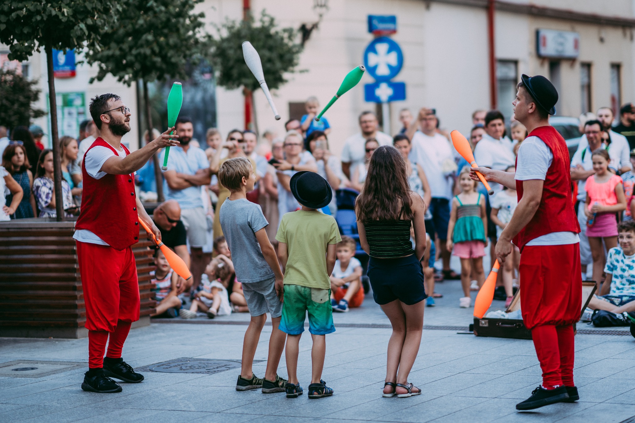Uliczny występ dwóch żonglerów w czerwonych strojach, żonglujących kolorowymi maczugami przed grupą dzieci i dorosłych na tle miejskiej ulicy.