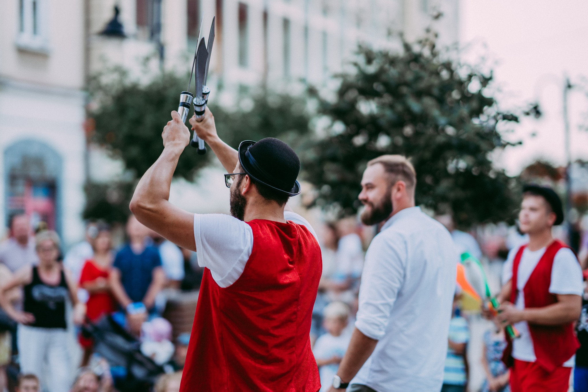 Mężczyzna w meloniku i czerwonej kamizelce prezentuje pokaz z dwoma mieczami skierowanymi ku górze, w tle publiczność i budynki.