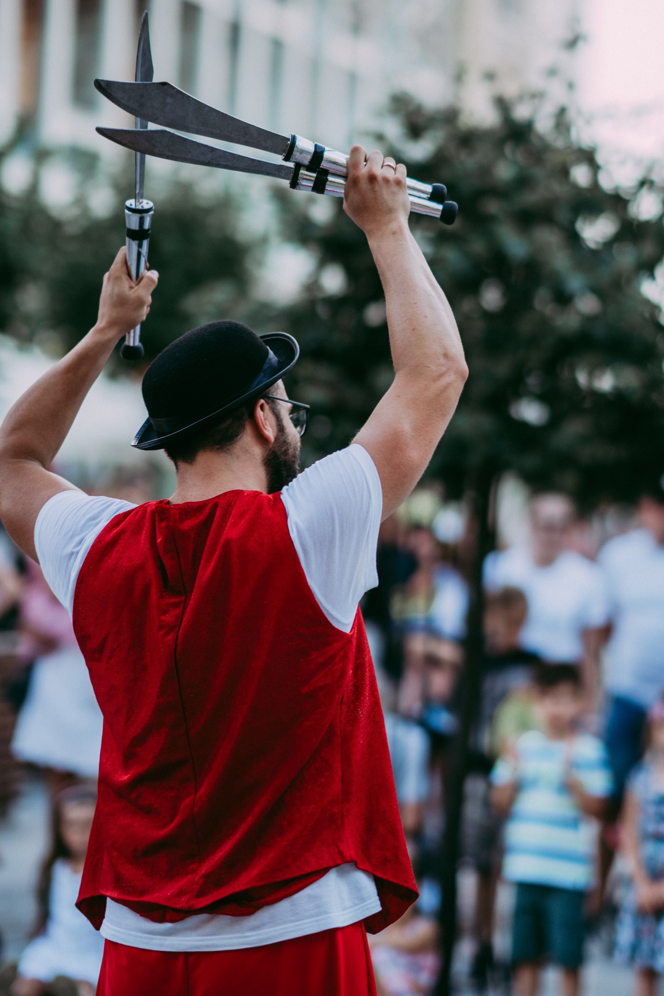 Iluzjonista w czerwonej kamizelce i meloniku prezentuje sztuczki z nożami na tle publiczności.