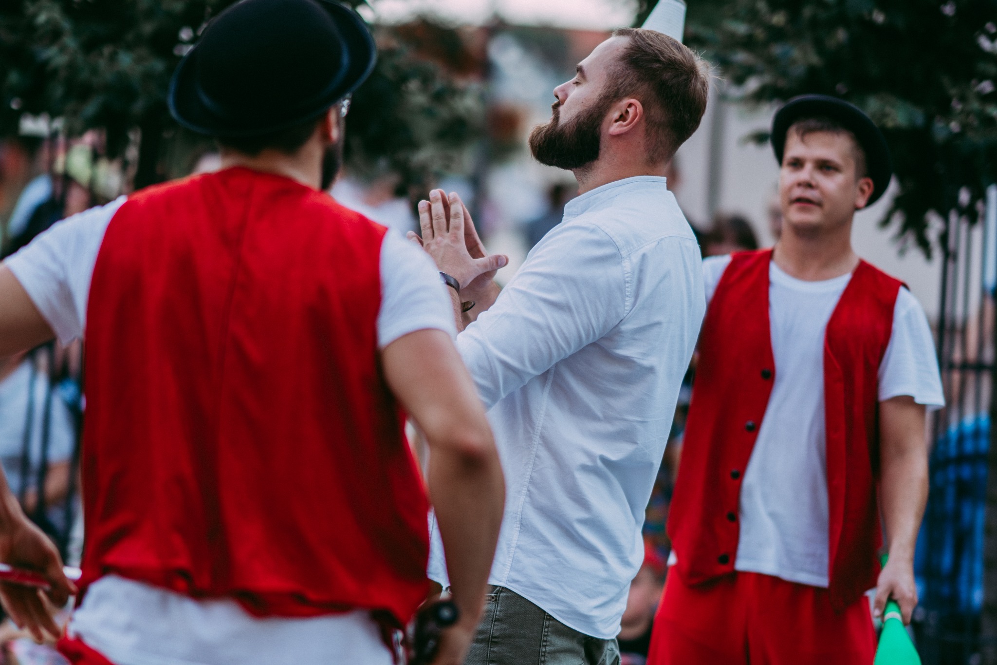 Trzech mężczyzn w kostiumach: dwaj w czerwonych kamizelkach i kapeluszach, jeden w białej koszuli, przygotowują się do występu ulicznego na tle drzew.
