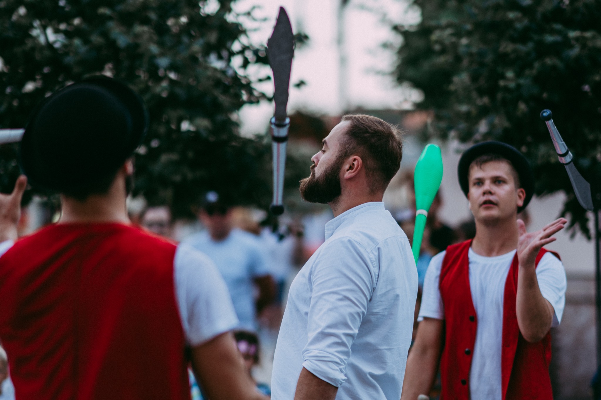 Mężczyzna z brodą w białej koszuli żongluje maczugami przed publicznością, drugi żongler w czerwonej kamizelce i kapeluszu w tle.