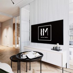 Łukasz Macuga architekt wnętrz - Wnętrze apartamentu z otwartą kuchnią, salonem i sypialnią. Parkiet w jodełkę, białe ściany w pionowe pasy, telewizor z logo architekta wnętrz.
