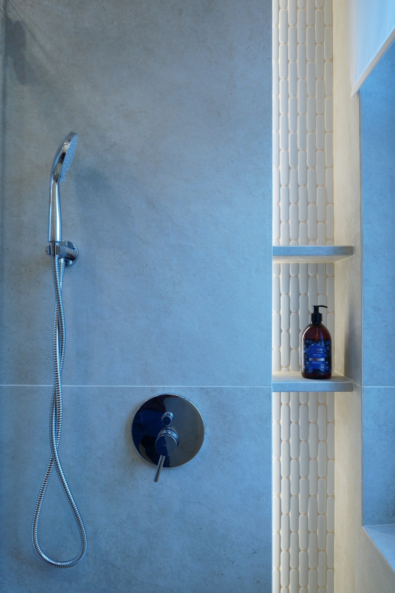 Nowoczesna łazienka z chromowaną słuchawką prysznicową, baterią i podświetlaną wnęką z półkami, na których stoi butelka mydła. Minimalistyczny design.