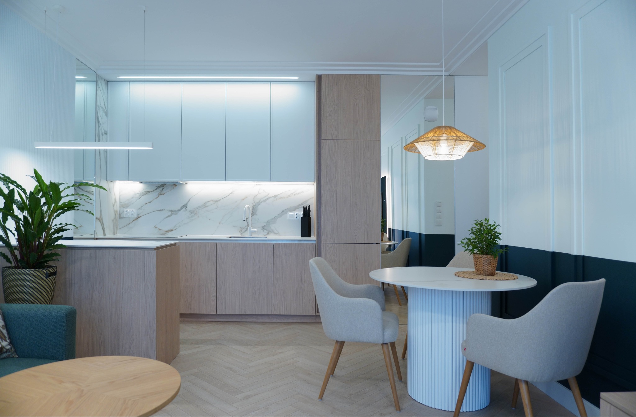 Jasna kuchnia z marmurowym blatem i drewnianymi szafkami, okrągły stół z krzesłami, oświetlenie LED i rośliny doniczkowe. Nowoczesny wystrój wnętrza.