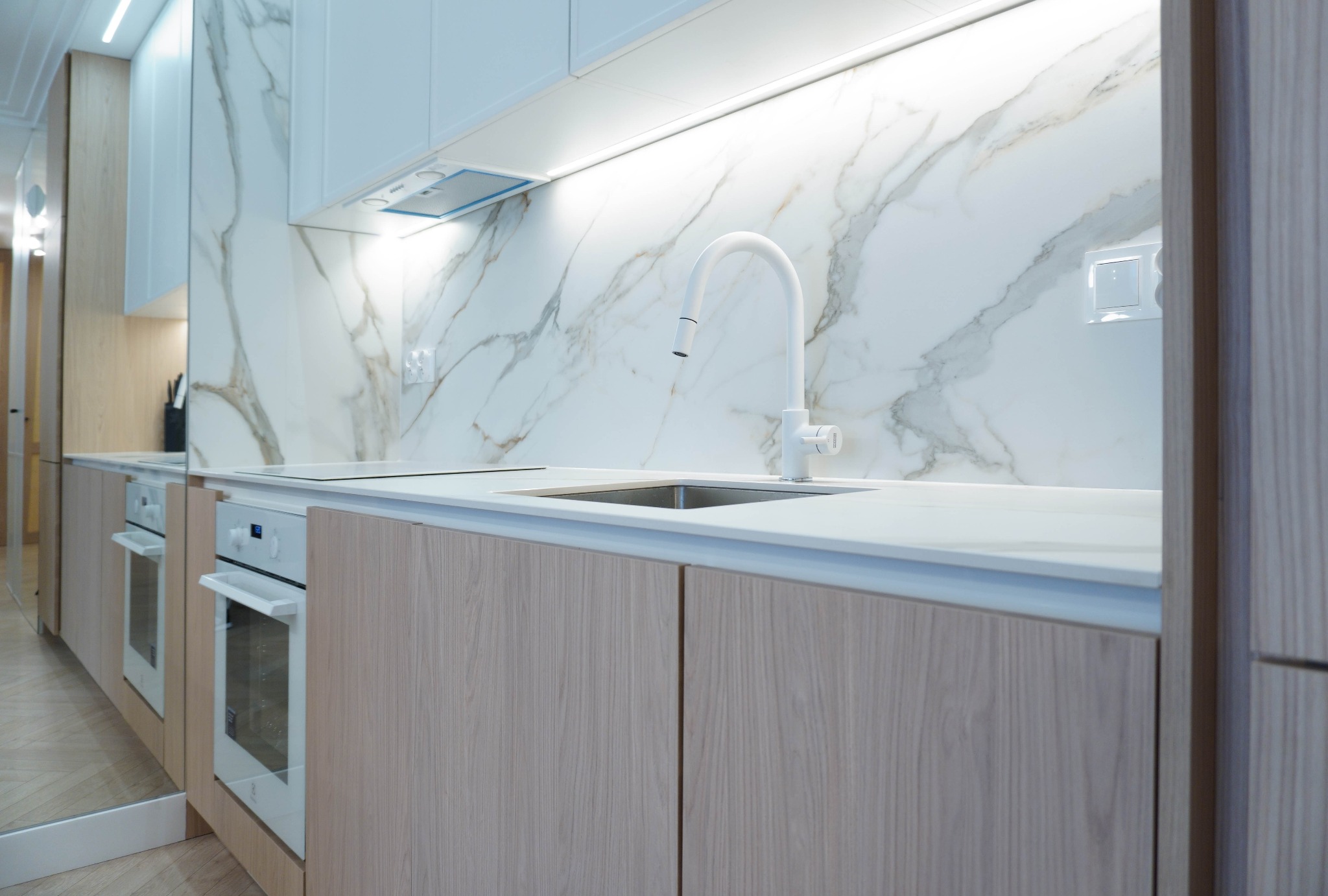 Jasna kuchnia z marmurowym backsplashiem i drewnianymi szafkami. Minimalistyczny design, biała armatura i oświetlenie LED. Ujęcie z niskiego kąta.