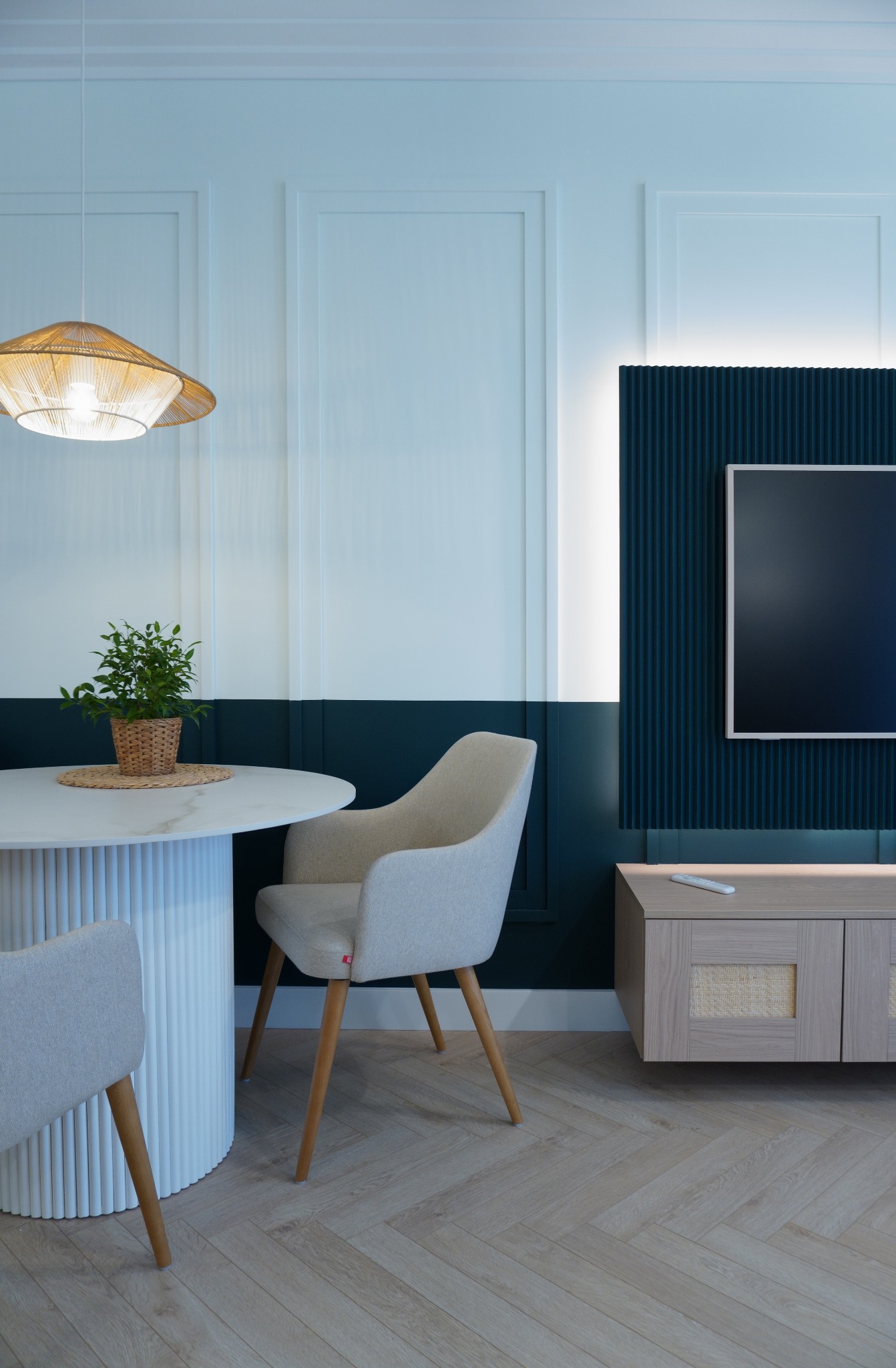 Nowoczesna jadalnia: okrągły stół z rośliną, krzesła z jasną tapicerką, ściana w dwóch odcieniach z panelem TV i szafką RTV. Parkiet ułożony w jodełkę.