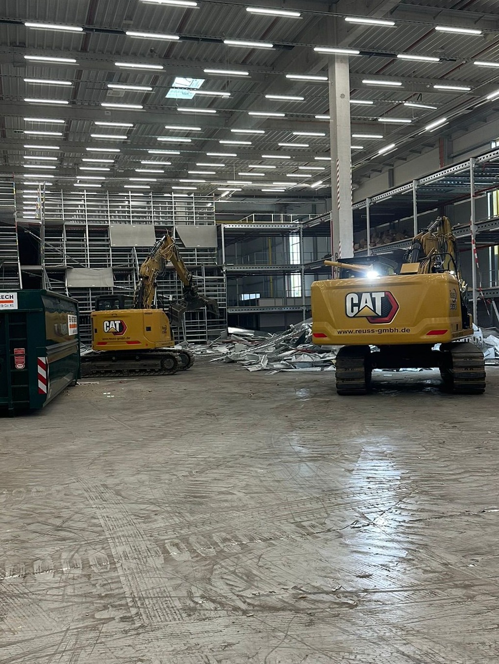 Dwa żółte, gąsienicowe, ciężkie  ekskawatory CAT w hali magazynowej w trakcie rozbiórki metalowej konstrukcji regałowej. Widoczny kontener na gruz.