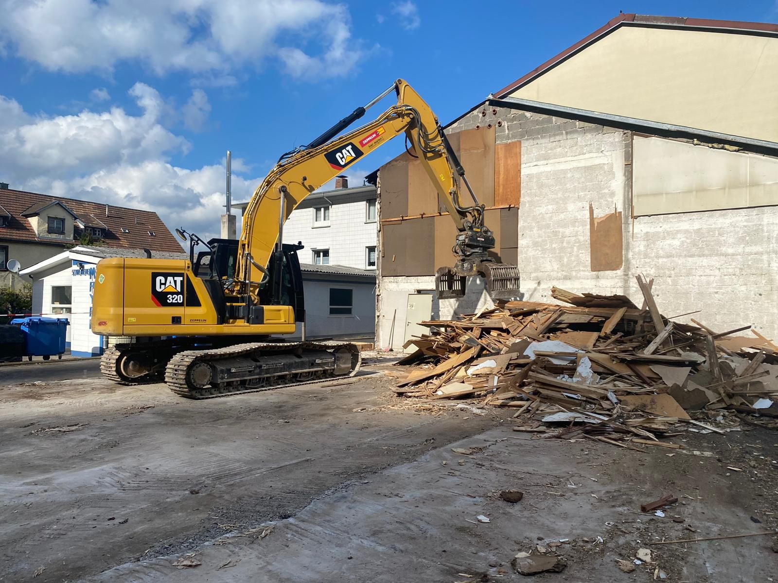 Żółta koparka CAT 320 z chwytakiem rozbiera budynek z szarej cegły. Wokół gruz i deski. Błękitne niebo z chmurami w tle. Częściowo widoczne budynki mieszkalne.