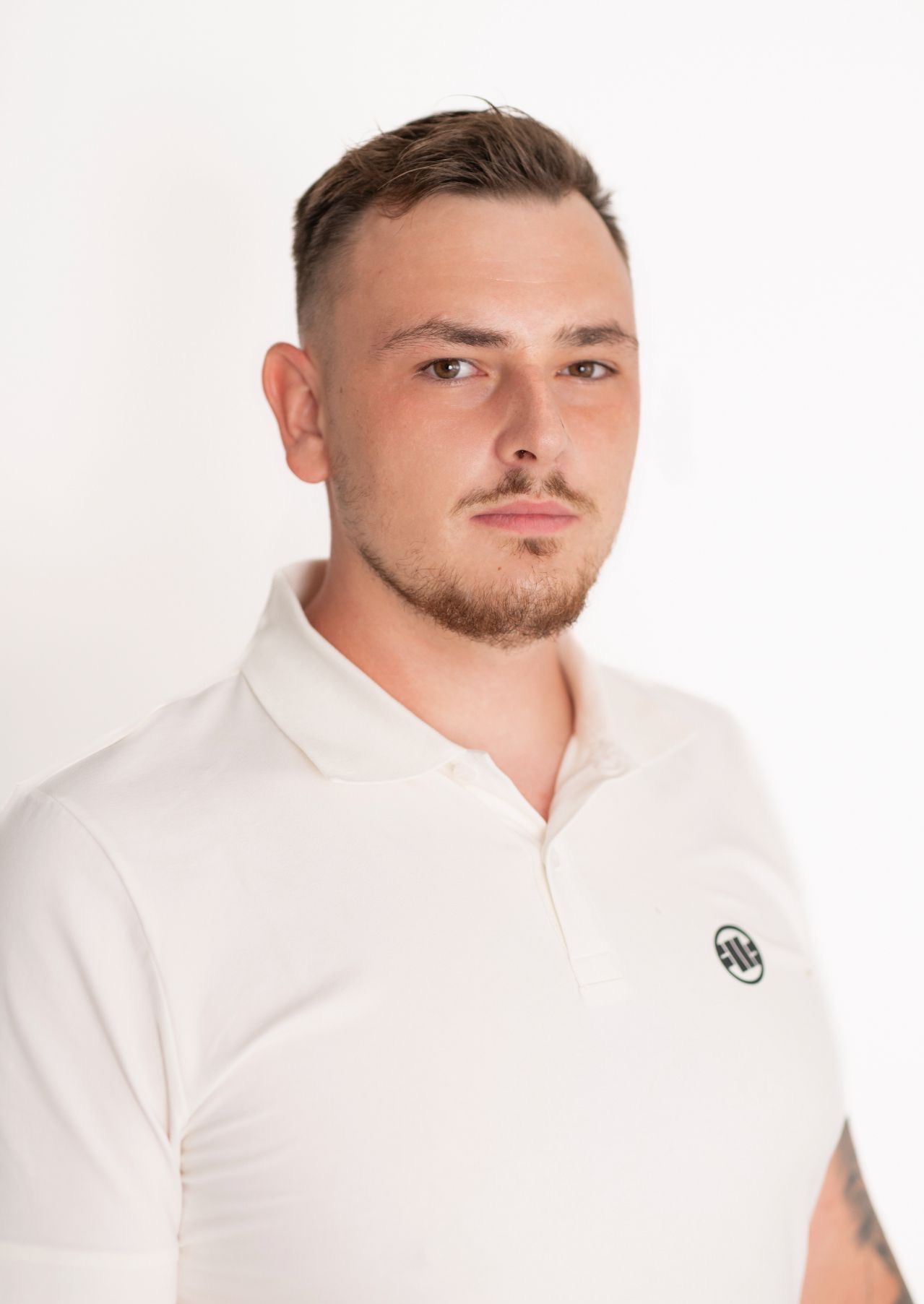Portret młodego mężczyzny z krótką brodą, ubranego w białą koszulkę polo z logo na tle jasnego studia. Ujęcie od przodu, skupione na twarzy i górnej części ciała.