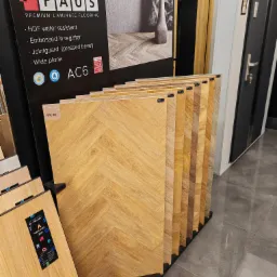 Ekspozycja paneli laminowanych marki FAUS z różnymi odcieniami drewna i wzorami, prezentowana w sklepie z drzwiami w tle. Widoczny szyld reklamowy z informacjami o właściwościach paneli, takich jak...
