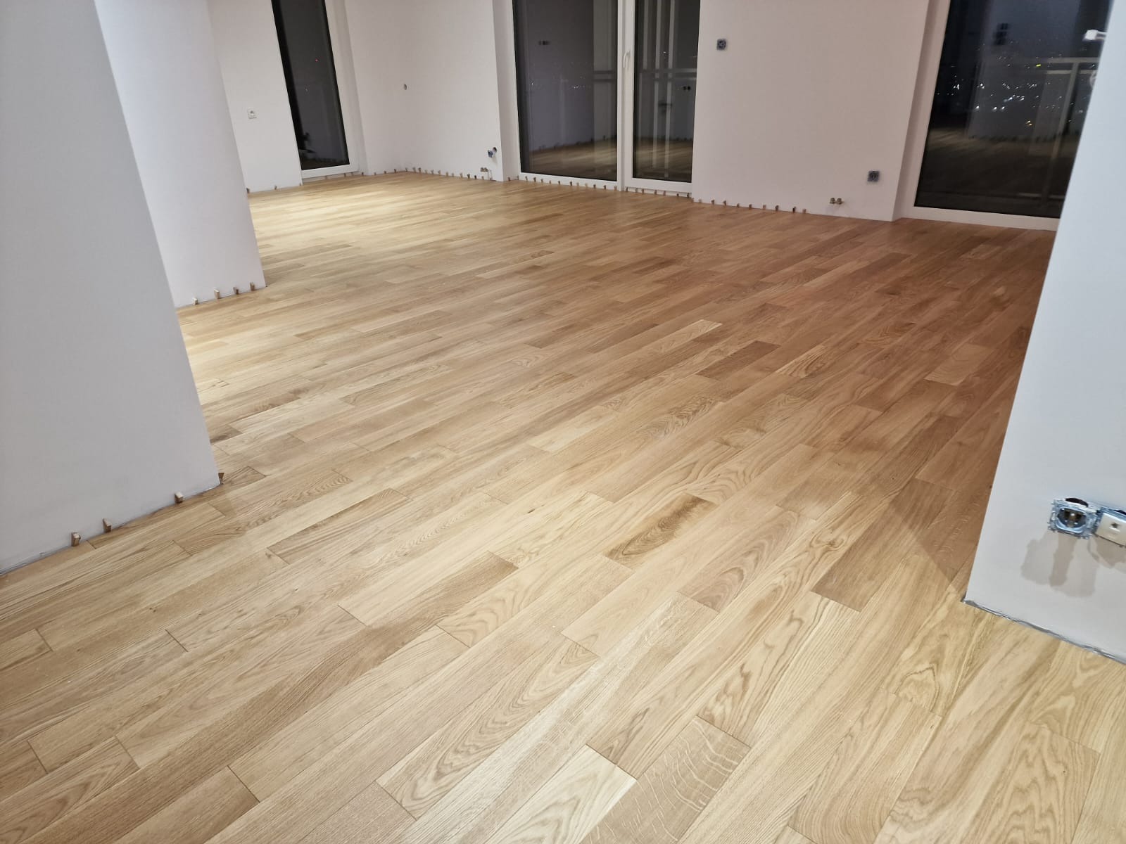 Jasne, drewniane panele podłogowe ułożone w przestronnym wnętrzu w Krakowie, widoczne listwy dystansowe przy ścianach i gniazdko elektryczne w dolnym rogu.