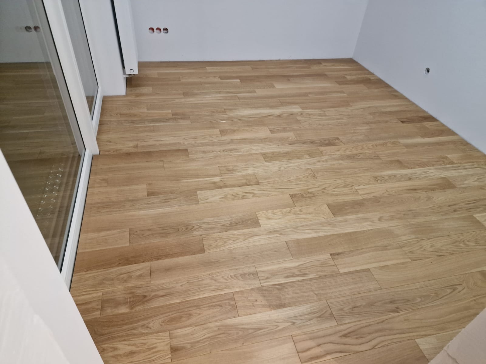 Ułożona podłoga z jasnych paneli drewnianych w pustym pokoju, widoczna dolna część okna balkonowego z białymi ramami.