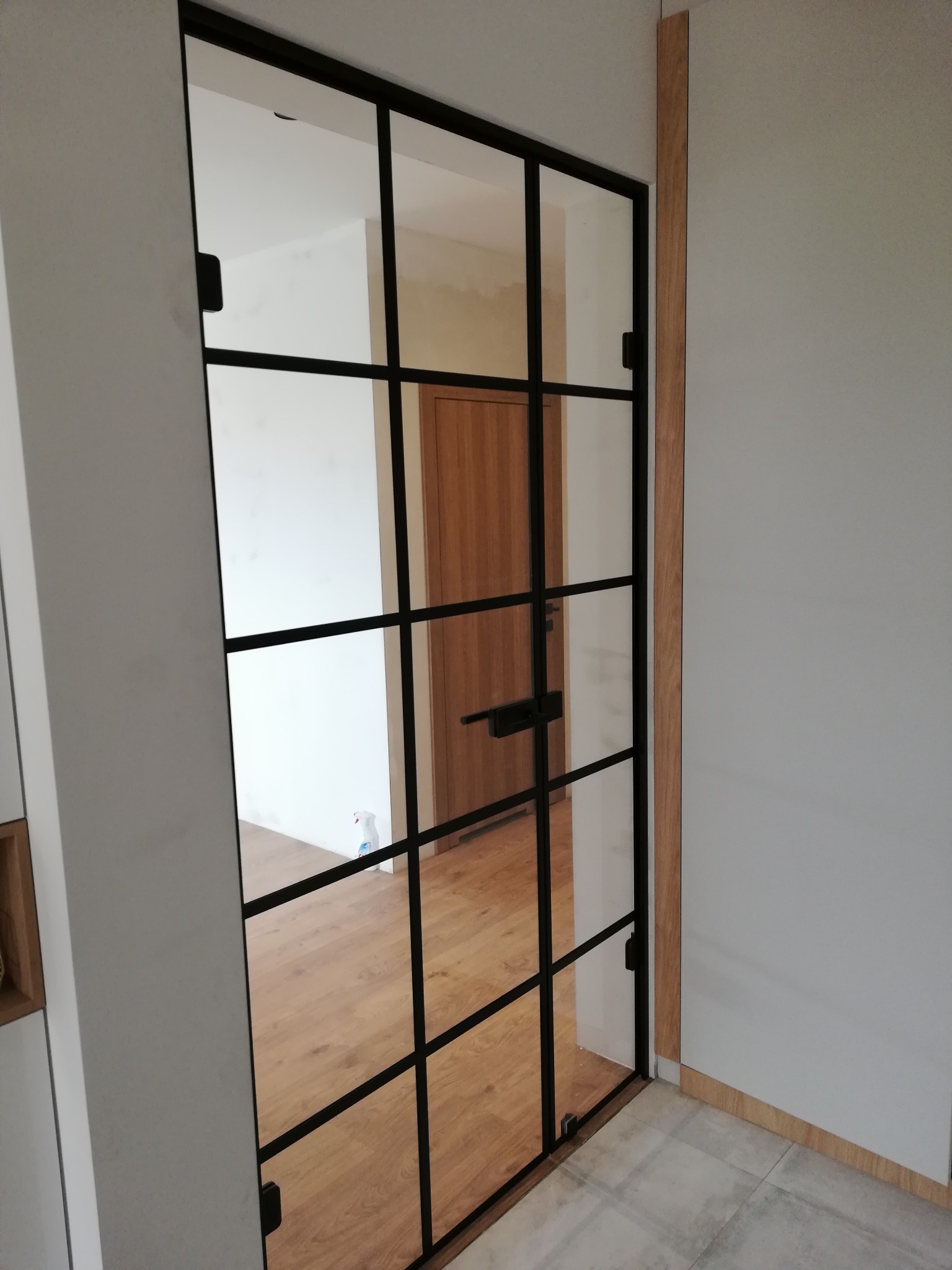 Szklane drzwi z czarnymi szprosami, widok na drewnianą podłogę i drzwi w tle, jasne ściany.