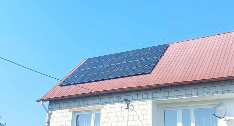 Panele słoneczne na dachu domu z cegły, widoczne okna i antena satelitarna, błękitne niebo w tle.