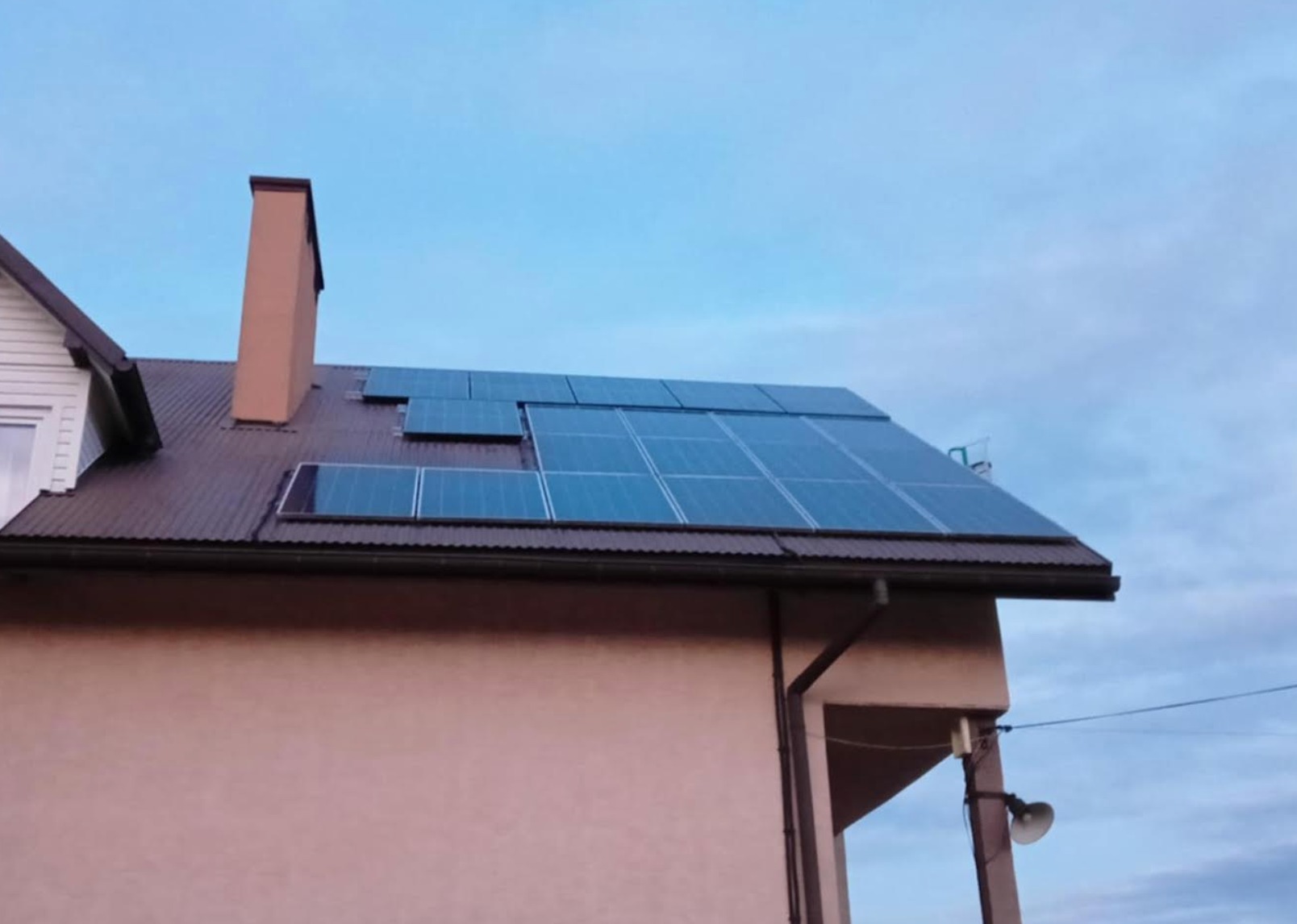 Instalacja paneli słonecznych na brązowym dachu domu jednorodzinnego z kominem; widok z dołu na panele o niebieskiej barwie, na tle błękitnego nieba.